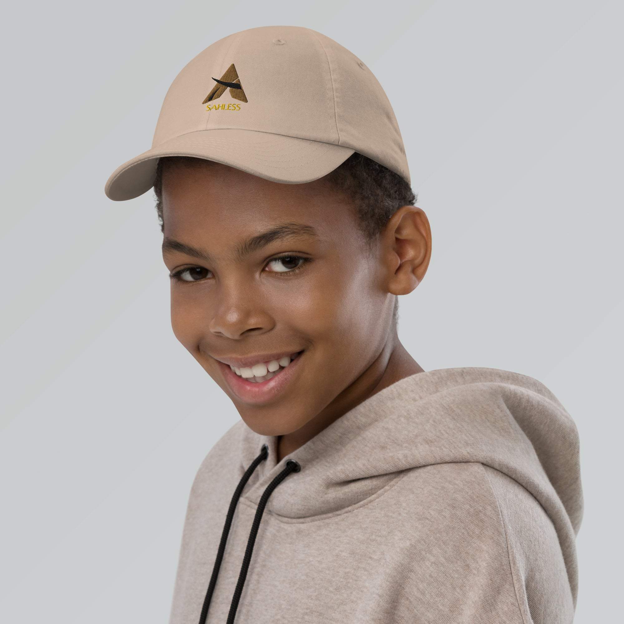 Youth wearing SahLess baseball cap, beige color, with adjustable strap and pre-curved visor for sun protection.